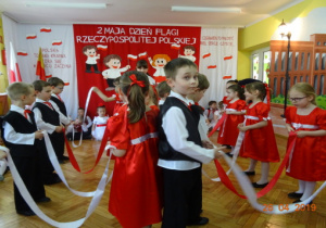 Dziewczynki w czerwonych sukienkach oraz chłopcy ubrani na galowo stoją w dwóch rzędach naprzeciwko siebie z białymi i czerwonymi wstążkami.